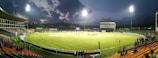 A brightly lit cricket stadium during an evening match with players on the field and spectators in the stands. The sky is partially clouded, adding a dramatic effect to the setting.
