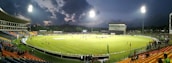 A brightly lit cricket stadium during an evening match with players on the field and spectators in the stands. The sky is partially clouded, adding a dramatic effect to the setting.