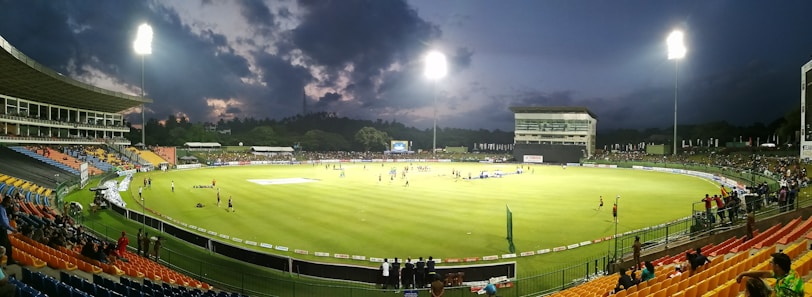 A brightly lit cricket stadium during an evening match with players on the field and spectators in the stands. The sky is partially clouded, adding a dramatic effect to the setting.