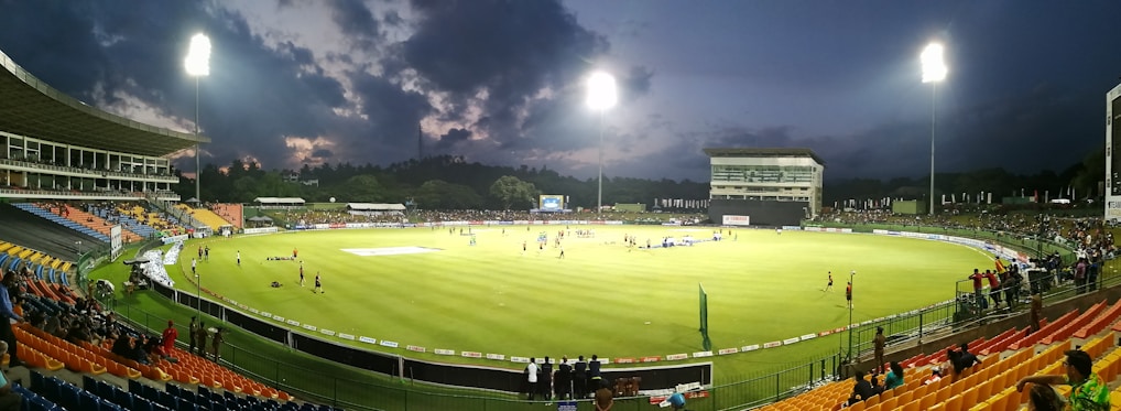 A brightly lit cricket stadium during an evening match with players on the field and spectators in the stands. The sky is partially clouded, adding a dramatic effect to the setting.