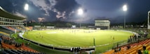 A brightly lit cricket stadium during an evening match with players on the field and spectators in the stands. The sky is partially clouded, adding a dramatic effect to the setting.