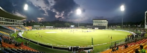 A brightly lit cricket stadium during an evening match with players on the field and spectators in the stands. The sky is partially clouded, adding a dramatic effect to the setting.