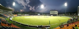 A brightly lit cricket stadium during an evening match with players on the field and spectators in the stands. The sky is partially clouded, adding a dramatic effect to the setting.