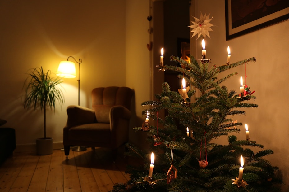 A cozy living room features a beautifully decorated Christmas tree adorned with candles and ornaments. A plush armchair sits in a corner near a potted plant, illuminated by a warmly glowing floor lamp. The ambiance is serene and festive, with soft lighting casting a comfortable glow.