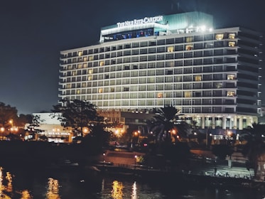 A large, well-lit hotel building with the name 'The Nile Ritz-Carlton' displayed prominently at the top. The structure features multiple floors with balconies, and the exterior is illuminated, giving it a grand appearance against the night sky. In the foreground, there are trees partially obscuring the building, and there are streetlights casting a warm glow. The image includes reflections in a nearby water surface, adding to the scene's elegance.