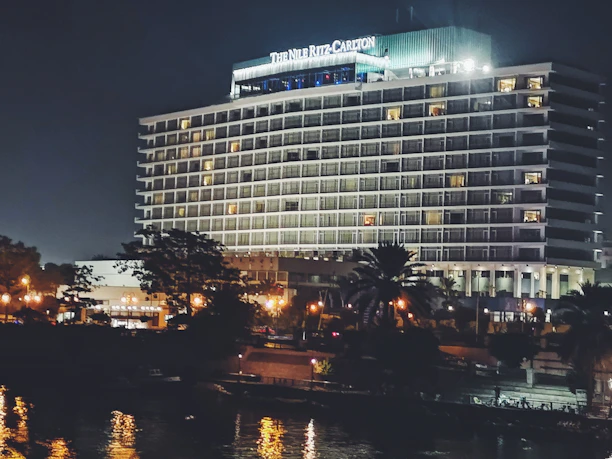 A large, well-lit hotel building with the name 'The Nile Ritz-Carlton' displayed prominently at the top. The structure features multiple floors with balconies, and the exterior is illuminated, giving it a grand appearance against the night sky. In the foreground, there are trees partially obscuring the building, and there are streetlights casting a warm glow. The image includes reflections in a nearby water surface, adding to the scene's elegance.