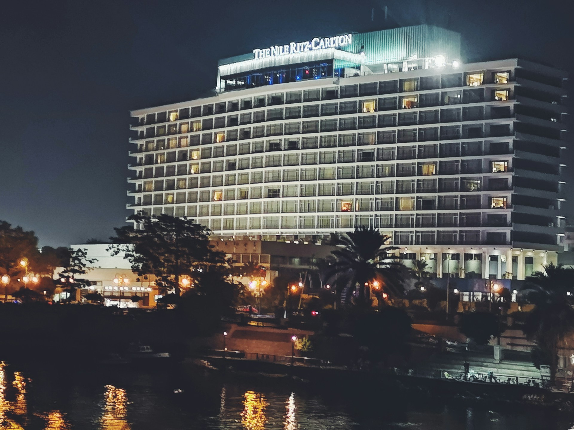 A large, well-lit hotel building with the name 'The Nile Ritz-Carlton' displayed prominently at the top. The structure features multiple floors with balconies, and the exterior is illuminated, giving it a grand appearance against the night sky. In the foreground, there are trees partially obscuring the building, and there are streetlights casting a warm glow. The image includes reflections in a nearby water surface, adding to the scene's elegance.