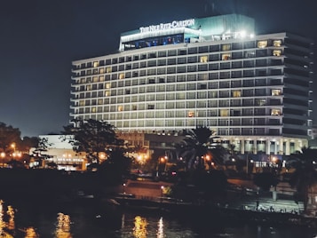 A large, well-lit hotel building with the name 'The Nile Ritz-Carlton' displayed prominently at the top. The structure features multiple floors with balconies, and the exterior is illuminated, giving it a grand appearance against the night sky. In the foreground, there are trees partially obscuring the building, and there are streetlights casting a warm glow. The image includes reflections in a nearby water surface, adding to the scene's elegance.