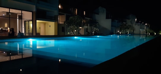 A serene nighttime scene featuring an illuminated swimming pool that reflects the soft glow of nearby lights. The setting appears to be a tranquil outdoor area adjacent to a modern building with large windows, possibly a hotel or resort. Subtle ambient lighting and surrounding trees contribute to the peaceful atmosphere.