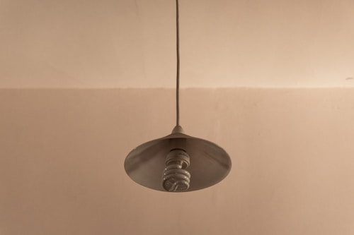 A single light fixture hangs from the ceiling, featuring a spiral-shaped, energy-efficient bulb under a simple, metallic lampshade. The background is a plain, light beige wall.