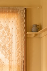 A delicate lace curtain with intricate floral patterns hangs beside a window. The curtain allows gentle, warm light to stream through, casting subtle, ornamental shadows. On the adjacent wall, a small, rustic blue pitcher sits on a wooden shelf, providing a touch of classic decor.
