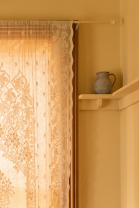 A delicate lace curtain with intricate floral patterns hangs beside a window. The curtain allows gentle, warm light to stream through, casting subtle, ornamental shadows. On the adjacent wall, a small, rustic blue pitcher sits on a wooden shelf, providing a touch of classic decor.