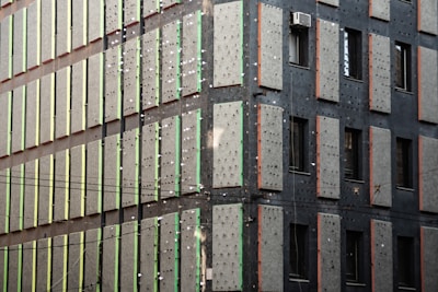 A building facade featuring a grid-like pattern with vertical rows of large, rectangular concrete slabs. Some of these slabs have small light-colored dots or holes. The building has an industrial appearance with air conditioning units protruding from some windows.