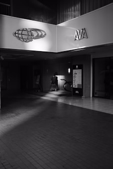 A dimly lit indoor space with tiled flooring features a person in the background, partially illuminated by light. There are signs displaying 'AVA' and a logo resembling a globe. A bicycle is parked near a wall, close to the person.