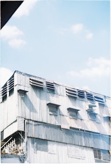 A large industrial building characterized by corrugated metal siding and small windows. The structure appears weathered, with visible rust and dirt stains. The sky above is bright with a few clouds, suggesting a sunny day.