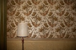 A vintage-style table lamp with a white shade stands against a wall adorned with ornate floral wallpaper. The wallpaper features a pattern of intricate green and pink flowers and vines. Below the wallpaper, there is a decorative textured paneling in a light beige color.