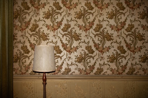A vintage-style table lamp with a white shade stands against a wall adorned with ornate floral wallpaper. The wallpaper features a pattern of intricate green and pink flowers and vines. Below the wallpaper, there is a decorative textured paneling in a light beige color.