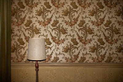 A vintage-style table lamp with a white shade stands against a wall adorned with ornate floral wallpaper. The wallpaper features a pattern of intricate green and pink flowers and vines. Below the wallpaper, there is a decorative textured paneling in a light beige color.