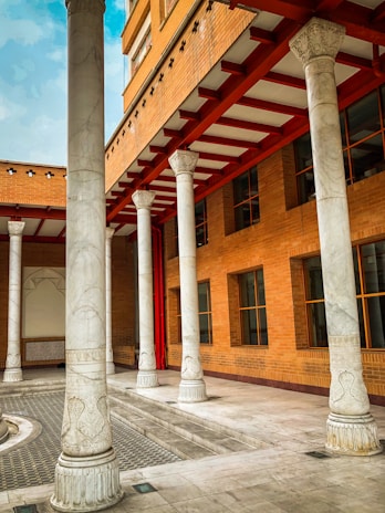 A series of tall, ornate marble columns support a building with a brick façade. The architecture features a combination of classical and modern elements. The red accents on the beams provide contrast against the predominantly neutral tones. Windows are symmetrically arranged along the brick wall.