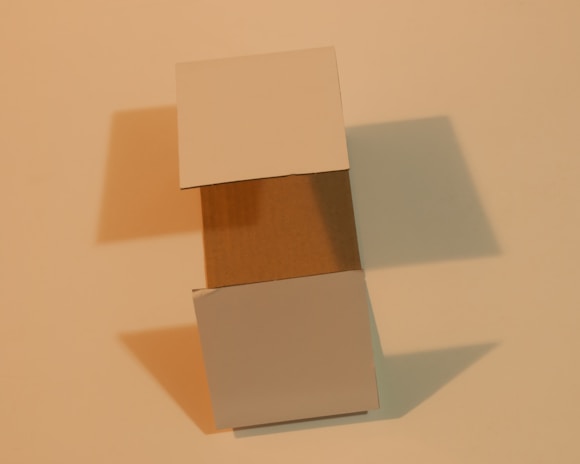 A three-dimensional cardboard box with its flaps open, casting soft shadows on a neutral beige surface. The lighting creates a warm tone, highlighting the texture of the cardboard.
