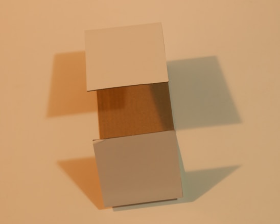 A three-dimensional cardboard box with its flaps open, casting soft shadows on a neutral beige surface. The lighting creates a warm tone, highlighting the texture of the cardboard.