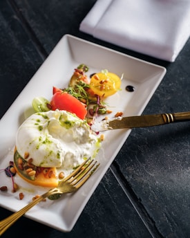 A neatly arranged gourmet dish on a rectangular white plate, featuring a creamy cheese, possibly burrata, placed on top of slices of colorful tomatoes, garnished with microgreens, pine nuts, and a drizzle of green sauce. The setting includes a gold fork and knife, with a folded white napkin in the background on a dark surface.