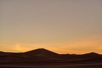 A serene desert landscape with gently rolling sand dunes under a gradient sky transitioning from soft orange to pale gray as the sun sets.