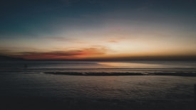The image captures a tranquil beach scene at sunset. The sky is adorned with a gradient of colors ranging from deep blue at the top to warm oranges and reds near the horizon. The calm ocean reflects these colors, and dark silhouettes of land and water create a serene atmosphere.
