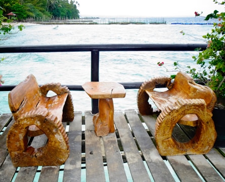 Two rustic wooden chairs and a small wooden table are placed on a balcony or deck overlooking the ocean. The deck has a wooden railing, and the chairs and table have a unique, handcrafted design with a natural finish. The background features a stretch of calm blue water and a distant shoreline with lush green trees and some structures.