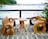 Two rustic wooden chairs and a small wooden table are placed on a balcony or deck overlooking the ocean. The deck has a wooden railing, and the chairs and table have a unique, handcrafted design with a natural finish. The background features a stretch of calm blue water and a distant shoreline with lush green trees and some structures.