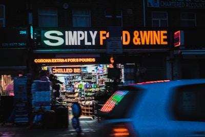 A brightly lit convenience store with a glowing sign that reads 'SIMPLY FOOD & WINE'. The storefront is lined with various products visible through the glass, and a neon sign advertises 'CORONA EXTRA 4 FOR 5 POUNDS'. A car is passing by in the foreground, slightly blurred due to motion.