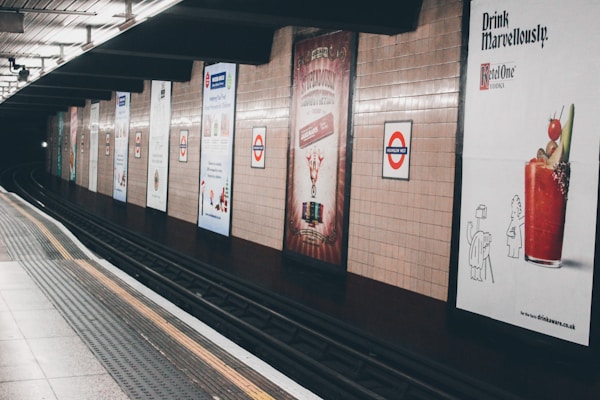 A subway station platform is lined with a series of advertisements on the tiled wall. The tracks curve away into the distance, creating a sense of depth. One advertisement prominently displays a cocktail, while others show a variety of designs and colors. The lighting is subdued, giving the station a quiet atmosphere.