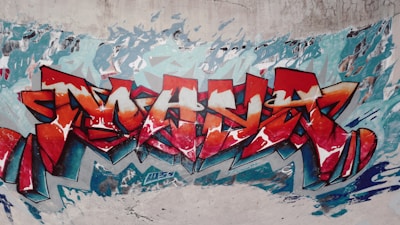A vivid and intricate graffiti mural features abstract shapes and bold lettering in shades of red, orange, and blue. The artwork covers a textured concrete wall, with dynamic strokes and patterns creating a sense of motion.