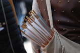 A quiver filled with numerous wooden arrows, each featuring a white tail feather. The person carrying the quiver is wearing a leather garment, likely a protective vest, decorated with small brass studs.