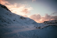 A snow-covered mountainous landscape under a sky with a mixture of soft clouds and the warm hues of sunset. A group of people can be seen walking along a path that winds between the mountains.