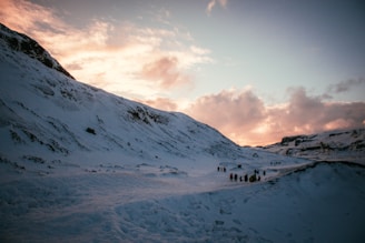 A snow-covered mountainous landscape under a sky with a mixture of soft clouds and the warm hues of sunset. A group of people can be seen walking along a path that winds between the mountains.