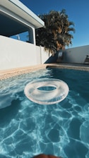 A clear swimming pool with a transparent inflatable ring floating on the water, framed by a light beige tiled border. In the background, a white building with a shaded patio and a wooden chair can be seen. Tall palm trees rise against a deep blue sky, enhancing the warm and sunny atmosphere.