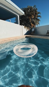 A clear swimming pool with a transparent inflatable ring floating on the water, framed by a light beige tiled border. In the background, a white building with a shaded patio and a wooden chair can be seen. Tall palm trees rise against a deep blue sky, enhancing the warm and sunny atmosphere.