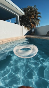 A clear swimming pool with a transparent inflatable ring floating on the water, framed by a light beige tiled border. In the background, a white building with a shaded patio and a wooden chair can be seen. Tall palm trees rise against a deep blue sky, enhancing the warm and sunny atmosphere.