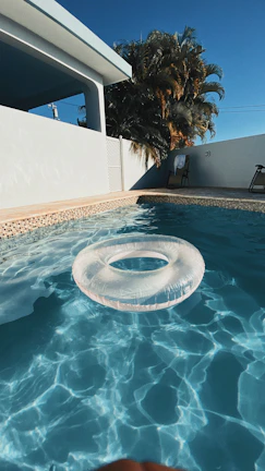 A clear swimming pool with a transparent inflatable ring floating on the water, framed by a light beige tiled border. In the background, a white building with a shaded patio and a wooden chair can be seen. Tall palm trees rise against a deep blue sky, enhancing the warm and sunny atmosphere.