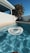 A clear swimming pool with a transparent inflatable ring floating on the water, framed by a light beige tiled border. In the background, a white building with a shaded patio and a wooden chair can be seen. Tall palm trees rise against a deep blue sky, enhancing the warm and sunny atmosphere.