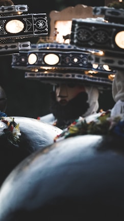 A close-up view of intricately designed hats, adorned with patterns and illuminated by small round lights. The hats appear to be part of a traditional costume, with dark fabric and floral decorations. A partially visible figure wearing one of the hats adds a mysterious element to the scene.