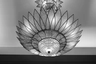 An ornate chandelier with a geometric, petal-like design composed of glass and metal elements. The intricate pattern resembles a large flower with layered sections. The central part features crystal-like spheres enhancing its luxurious appearance.