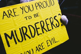 A bright yellow protest sign with bold black letters reads 'ARE YOU PROUD TO BE MURDERERS?' and 'YOU ARE EVIL.' A hand, wearing a glove, is holding the right edge of the sign.