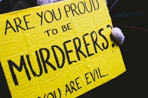 A bright yellow protest sign with bold black letters reads 'ARE YOU PROUD TO BE MURDERERS?' and 'YOU ARE EVIL.' A hand, wearing a glove, is holding the right edge of the sign.