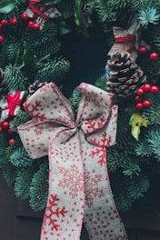 A decorative wreath with lush green pine branches, adorned with pinecones, red berries, and a large bow featuring red snowflake patterns on beige fabric. The wreath's natural elements are complemented by small red ribbons tied around some of the foliage, adding to the festive appearance.