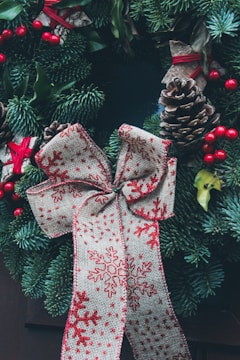 A decorative wreath with lush green pine branches, adorned with pinecones, red berries, and a large bow featuring red snowflake patterns on beige fabric. The wreath's natural elements are complemented by small red ribbons tied around some of the foliage, adding to the festive appearance.