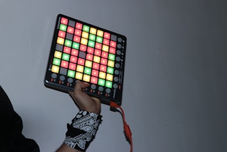 A person holds a device featuring a grid of colorful, illuminated buttons with a distinctive, patterned wristband on one wrist. The buttons are lit in various colors including red, green, and yellow, with a red cable extending from the device.
