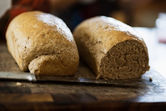 Two loaves of bread, one partially sliced, sit on a wooden cutting board next to a knife. The bread has a brown, rustic crust and a dense, soft interior, suggesting it is freshly baked. The background is blurred, focusing attention on the bread.