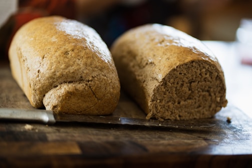 Two loaves of bread, one partially sliced, sit on a wooden cutting board next to a knife. The bread has a brown, rustic crust and a dense, soft interior, suggesting it is freshly baked. The background is blurred, focusing attention on the bread.
